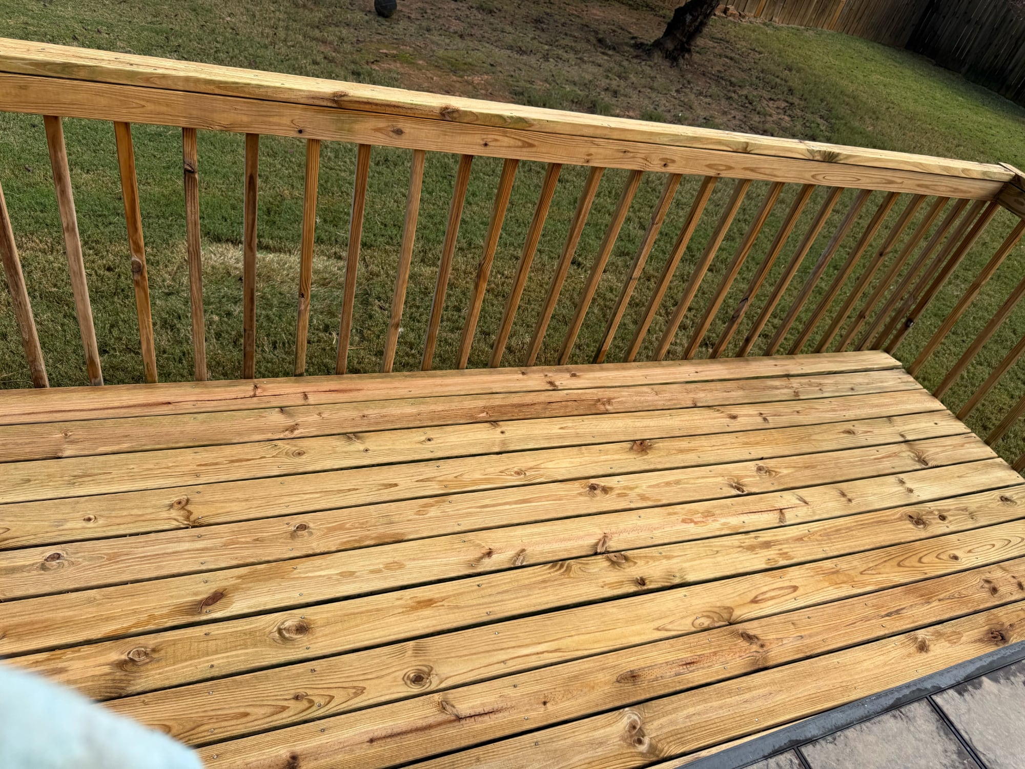 A wooden deck with a railing, showing natural wood grain and color, overlooking a grassy area.
