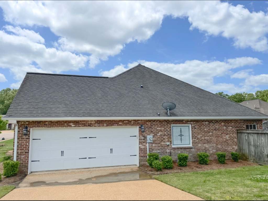 Front view of a brick house with a two-car garage, satellite dish, and landscaped garden under a blue sky with clouds.