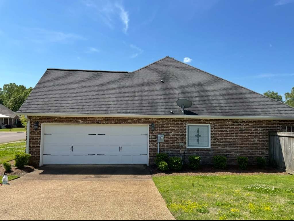 Front view of a brick house with a two-car garage, a satellite dish on the roof, and green shrubs in the yard under a clear blue sky.
